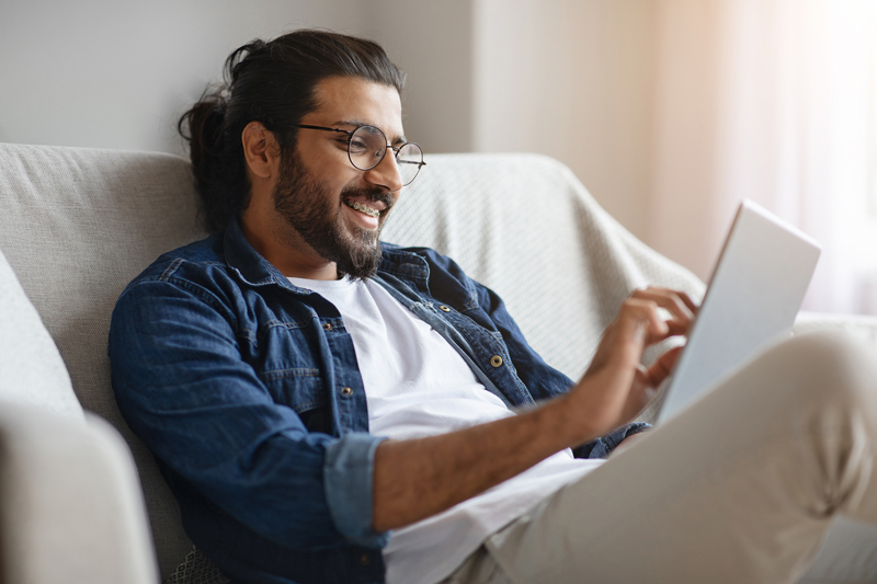 A man with glasses and a beard, wearing a denim shirt and white t-shirt, sits on a sofa smiling while using a laptop. - Raleigh Orthodontics